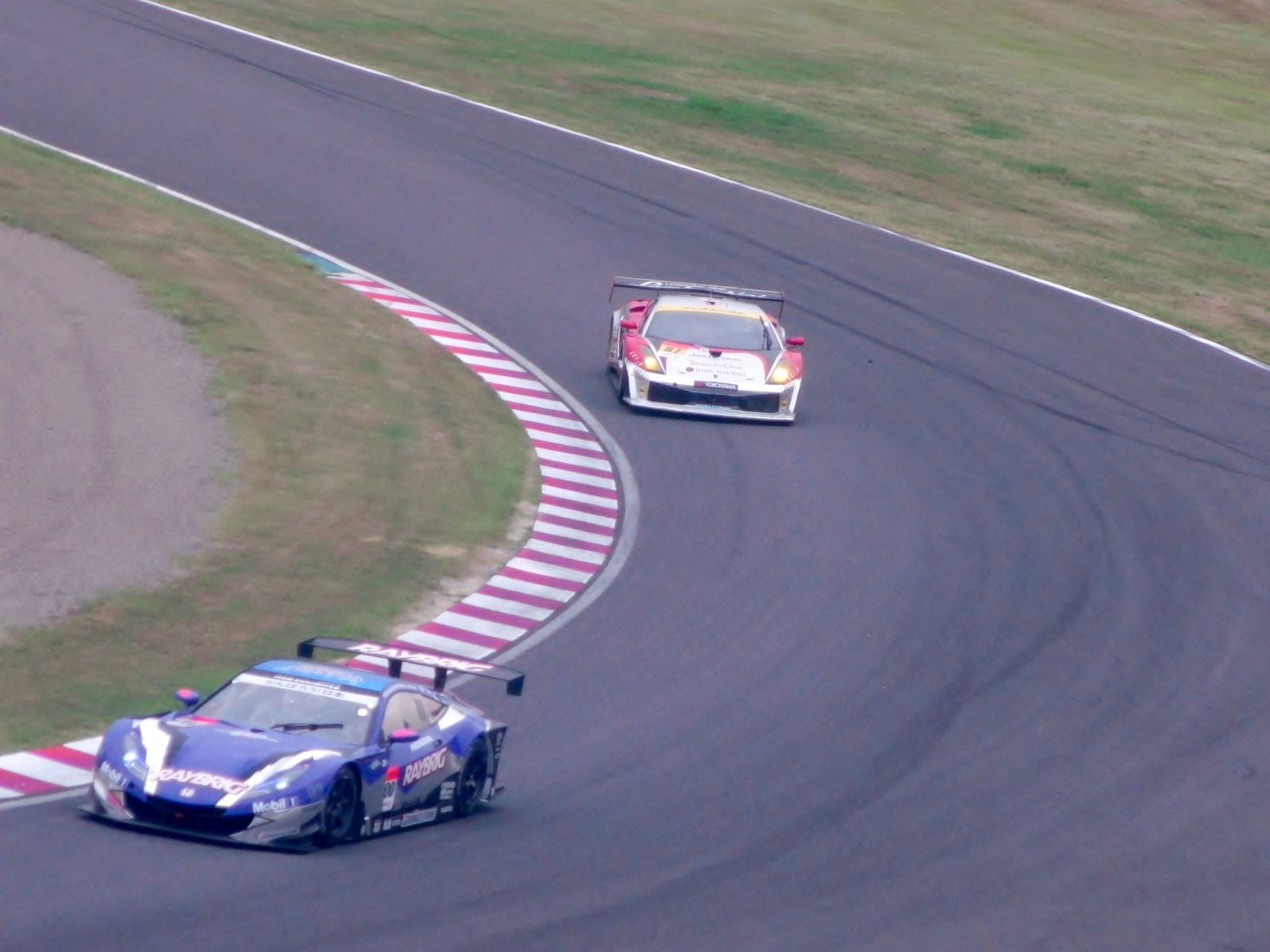 Zwei bunt lackierte Sportwagen auf einer kurvenreichen Strecke, wobei einer leicht vorne ist, umgeben von Gras.