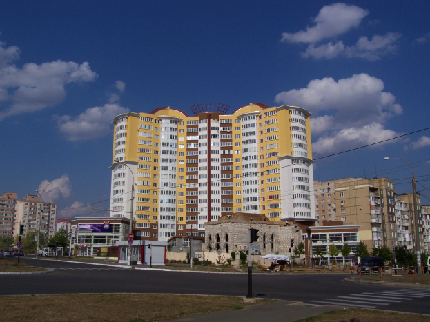 Ein großes Wohngebäude in einer Stadt umgeben von Straßenlaternen, Strommasten, Stromkabeln, Kraftfahrzeugen, Infotafeln, Bäumen und einer bewölkten Himmel
