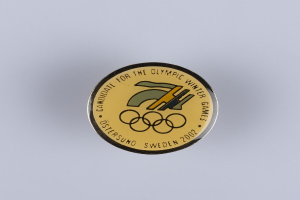 Ein schwarzes Kreisabzeichen mit einem goldenen Olympischen Winterspiellogo und weißer Schrift auf einer weißen Fläche.