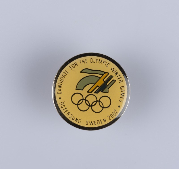 Ein schwarzes Kreisabzeichen mit einem goldenen Olympischen Winterspiellogo und weißer Schrift auf einer weißen Fläche.