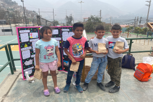 Eine Gruppe von Kindern steht auf einem Basketballfeld, jedes hält einen Papiertüte, mit Tüten und Gegenständen rechts und einer Texttafel links, mit Zaun, Pfosten, Straßenlaternen, Strommasten mit Drähten, Bäumen, Gebäuden, Hügeln und Himmel im Hintergrund.