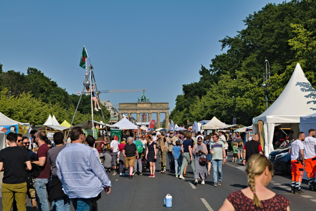 Eine Menschenmenge, die eine von Zelten, Fahrzeugen und Bäumen gesäumte Straße entlanggeht, mit einem Bogen und einem klaren blauen Himmel im Hintergrund und Fahnenmasten auf der linken Seite, wahrscheinlich zeigt das Oktoberfest in München, Deutschland.