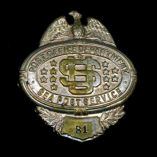 Ein silbern glänzender Abzeichen mit der Aufschrift 'Post Office Department Sea Post Service' auf schwarzem Hintergrund.
