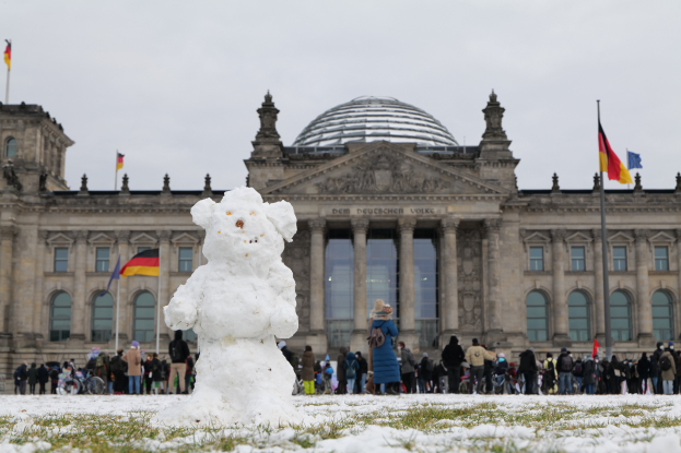 Ein Schneemann steht vor dem Reichstaggeb├Ąude in Berlin, Deutschland, umgeben von Menschen, einige halten Taschen, mit den Fenstern, Pfeilern, Bogengängen und Fahnenmasten des Geb├Ąudes im Hintergrund, unter einem klaren Himmel.