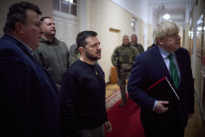 Boris Johnson, der Premierminister des Vereinigten Königreichs, steht in einem Flur und hält ein Buch in der Hand, umgeben von einer Gruppe von Menschen, darunter einige in Militäruniformen, mit Türen und Fenstern im Hintergrund.