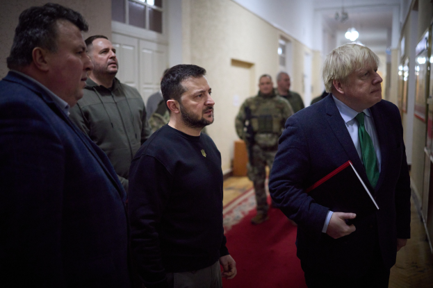 Boris Johnson, der Premierminister des Vereinigten Königreichs, steht in einem Flur und hält ein Buch in der Hand, umgeben von einer Gruppe von Menschen, darunter einige in Militäruniformen, mit Türen und Fenstern im Hintergrund.