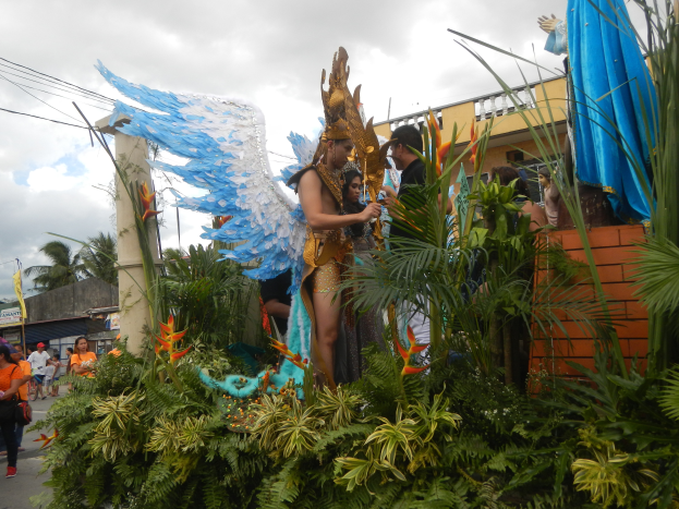 Eine Gruppe von Menschen geht neben einem Karnevalswagen mit Pflanzen und Blumen schmückungen, kostümierten Personen auf dem Wagen, Gebäuden, Bäumen und Strommasten mit Drähten im Hintergrund unter einem bewölkten Himmel.