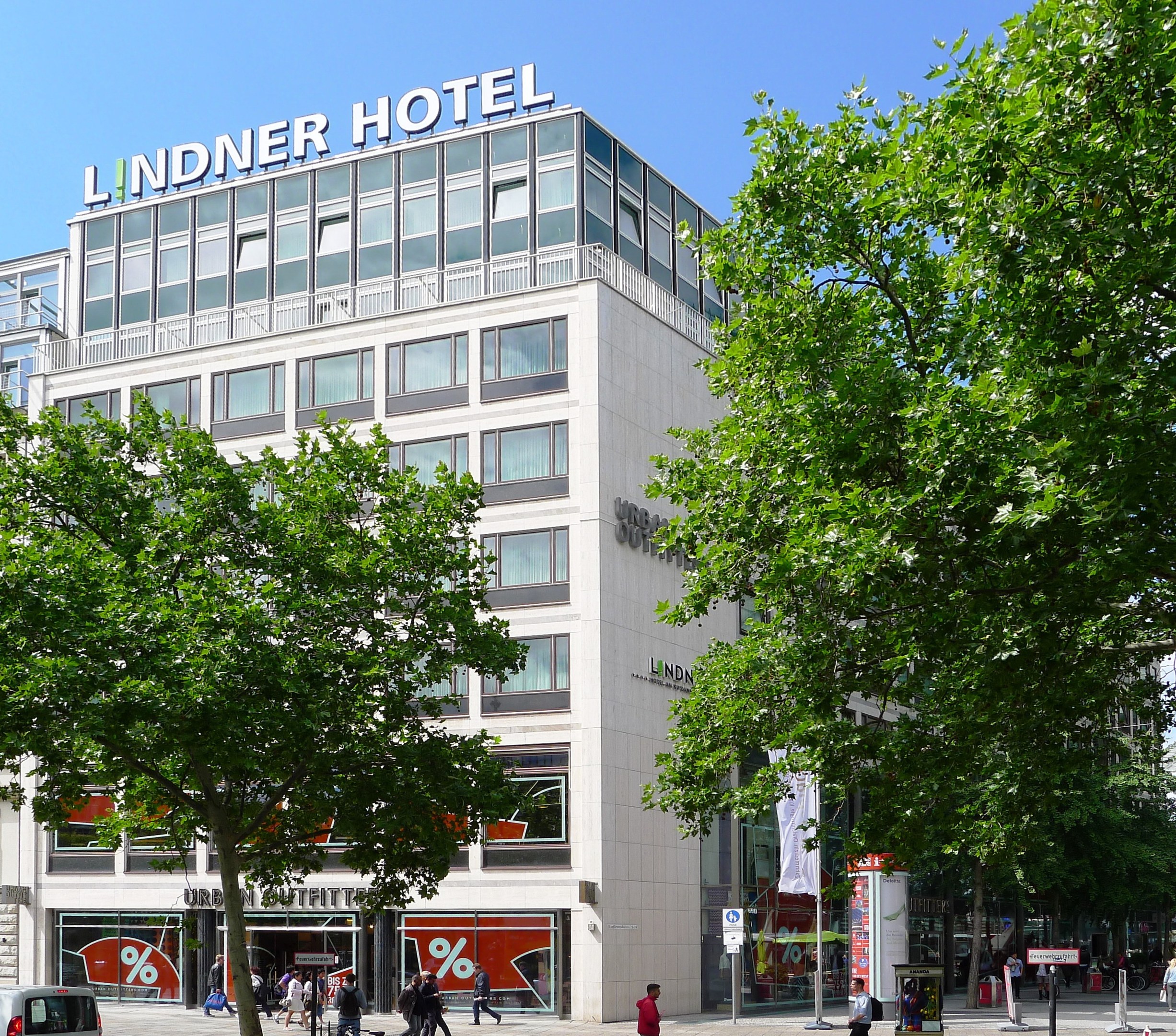 Großes Glasgebäude mit "Lindner Hotel"-Schriftzug oben, umgeben von Bäumen und einer belebten Straße mit Fußgängern und Fahrzeugen unter einem sichtbaren Himmel.