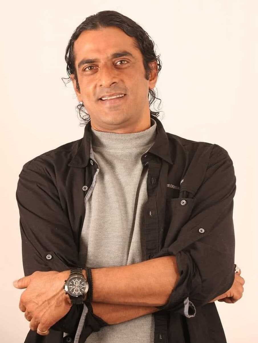 Ein Mann in einem schwarzen und aschfarbenen Kleid mit verschr├Ąckten Armen, der eine Uhr tr├Ągt und gegen einen wei├čen Hintergrund steht, der in Gedanken versunken zu sein scheint.