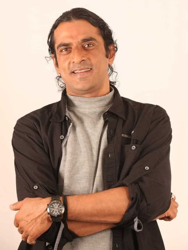 Ein Mann in einem schwarzen und aschfarbenen Kleid mit verschr├Ąckten Armen, der eine Uhr tr├Ągt und gegen einen wei├čen Hintergrund steht, der in Gedanken versunken zu sein scheint.