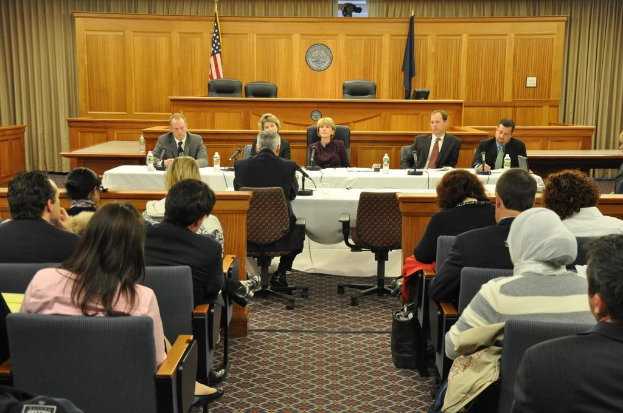 Ein Tagungsraum mit einer Gruppe von Menschen, zwei ausländischen Flaggen, einem orangen Schreibtisch mit Wasserflaschen und einem Mikrofon.