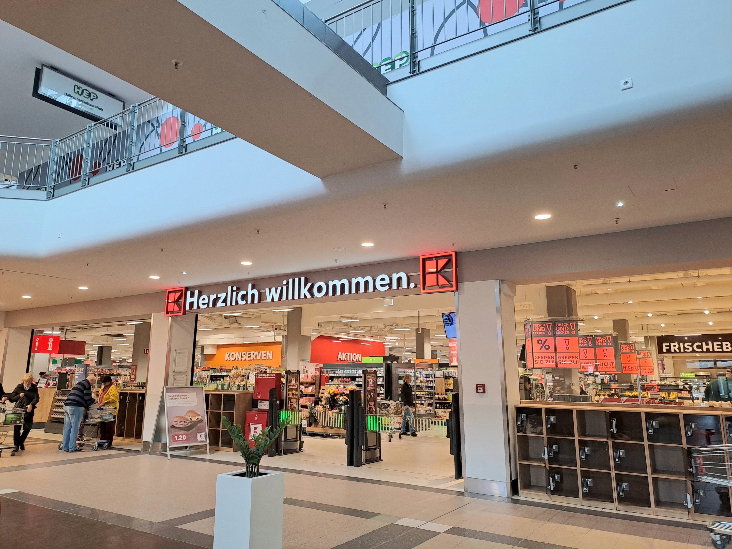 Innenansicht eines Einkaufszentrums mit Menschen, Geschäften, Infotafeln mit der Aufschrift "Herzlich willkommen", Beleuchtung, Geländern und einem Topfpfanzen am Boden.