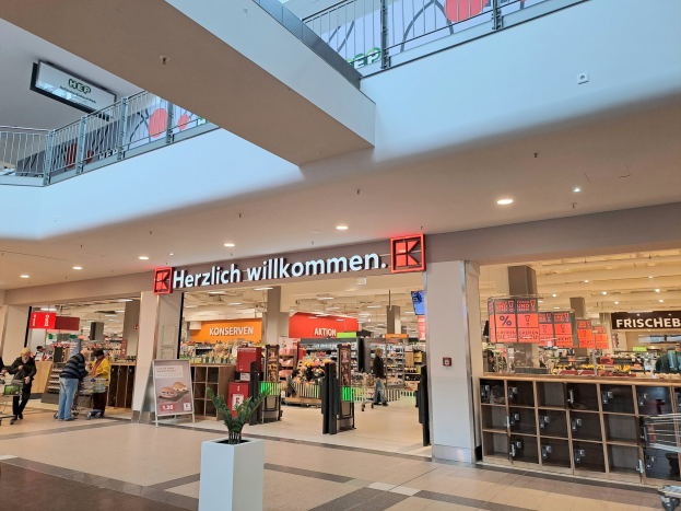 Innenansicht eines Einkaufszentrums mit Menschen, Geschäften, Infotafeln mit der Aufschrift "Herzlich willkommen", Beleuchtung, Geländern und einem Topfpfanzen am Boden.