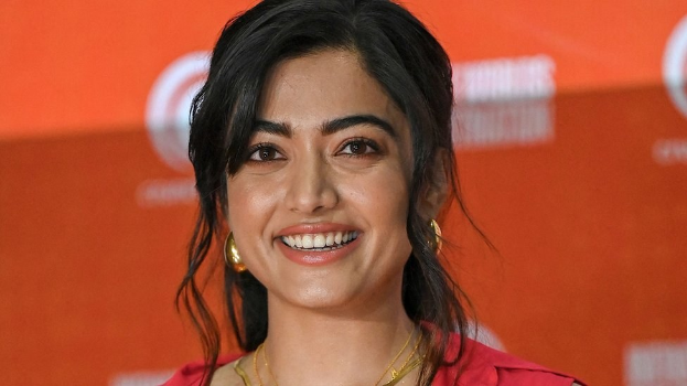 Eine Frau in einem roten Kleid mit einem eleganten Dutt und dezentem Make-up steht lächelnd da, trägt schwarze High Heels und ein gelbes Banner mit der Aufschrift 'Die besten Schönheits Looks von Sonam Kapoor beim Cannes Film Festival' im Hintergrund.