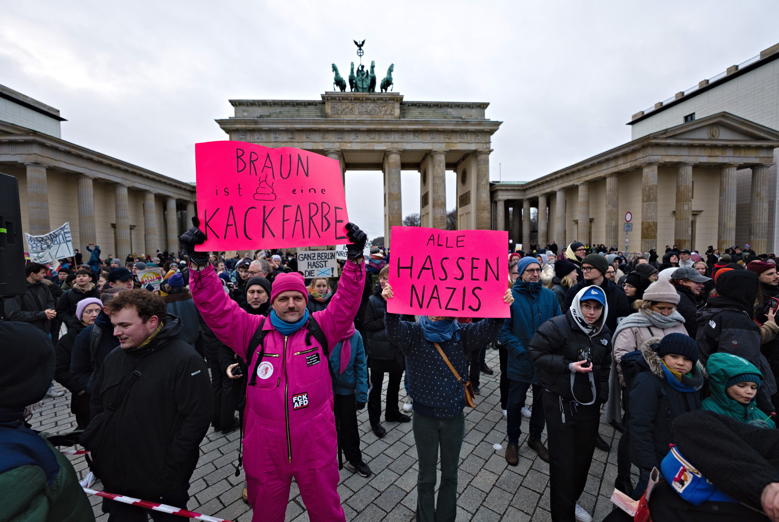 Eine Gruppe von Menschen protestiert vor dem Brandenburger Tor in Berlin, hält Schilder gegen die Anti-Nazis-Bewegung der deutschen Regierung und hat Gebäude und Statuen im Hintergrund unter einem klaren blauen Himmel.