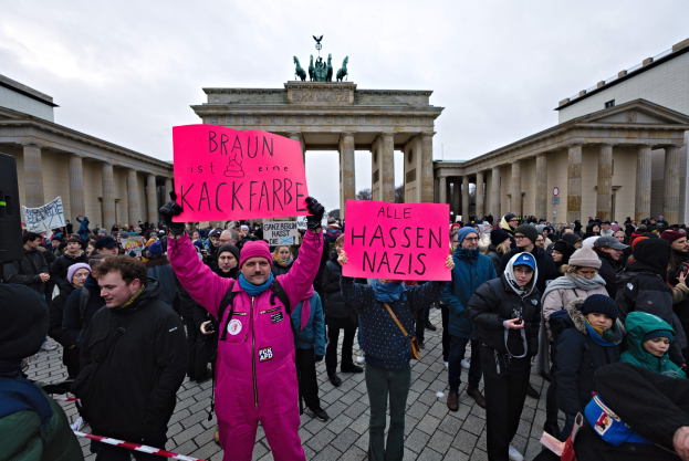 Eine Gruppe von Menschen protestiert vor dem Brandenburger Tor in Berlin, hält Schilder gegen die Anti-Nazis-Bewegung der deutschen Regierung und hat Gebäude und Statuen im Hintergrund unter einem klaren blauen Himmel.