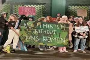 Eine Gruppe von Menschen hält ein Transparent mit der Aufschrift "Kein Feminismus ohne Trans-Frauen" und verschiedene Schilder in einem öffentlichen Raum mit einer Wand und einem Rollladen im Hintergrund.