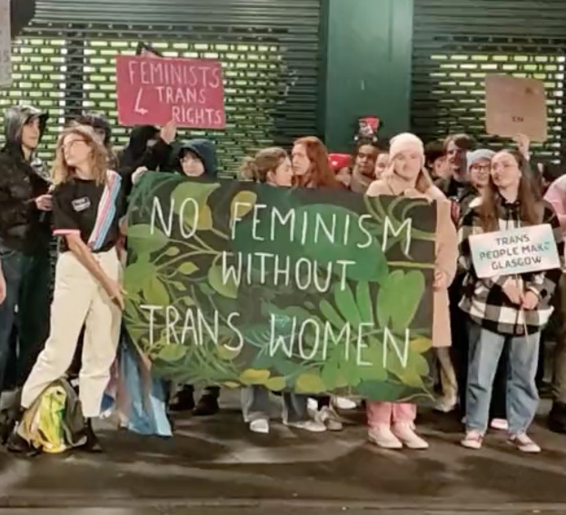 Eine Gruppe von Menschen hält ein Transparent mit der Aufschrift "Kein Feminismus ohne Trans-Frauen" und verschiedene Schilder in einem öffentlichen Raum mit einer Wand und einem Rollladen im Hintergrund.