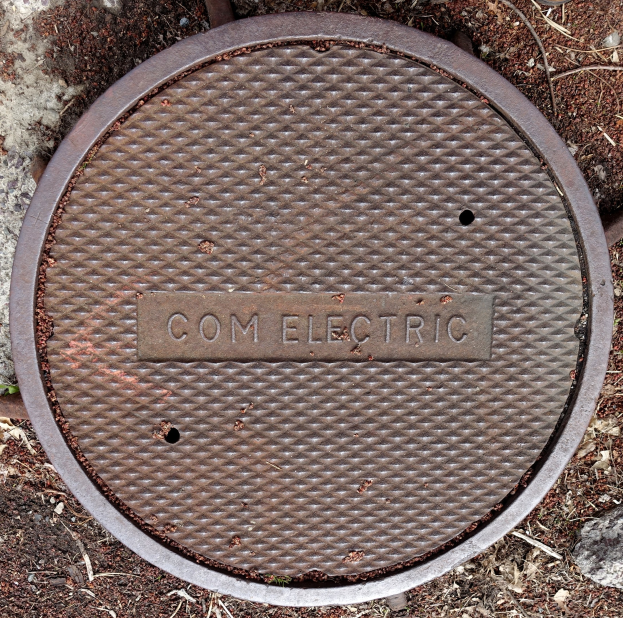 Ein Kanaldeckel auf dem Boden mit der Aufschrift "com electric", umgeben von Steinen und Zweigen.