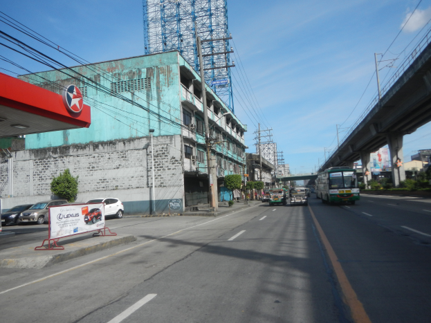 Eine Stadtstraße mit Autos und Bussen, einer Tankstelle links, Gebäuden, Strommasten mit Drähten, einer Brücke rechts, Bäumen und einem bewölkten Himmel.