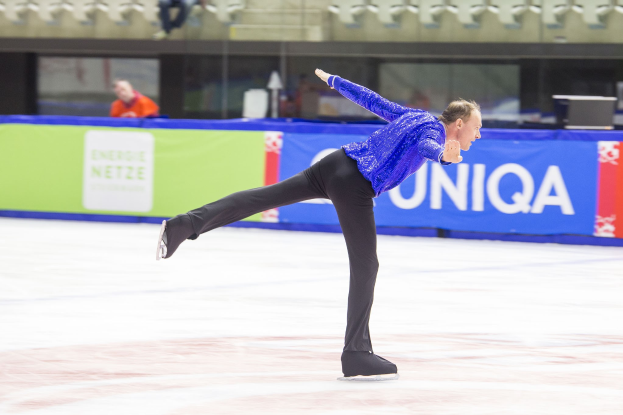Ein Mann in einem blauen Hemd und schwarzen Hosen, der auf einer Eisbahn während der World Figure Skating Championships skatet, mit einem Banner im Hintergrund, das "World Figure Skating Championships - Day 2" lautet, und einigen Zuschauern in den Zuschauerrängen.