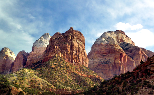 Eine malerische Aussicht auf den Zion-Nationalpark in Utah mit majestätischen Bergen, grünen Bäumen, steinigem Gelände und einem Himmel mit weißen, flauschigen Wolken.
