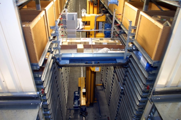 Großes Lagerinnere mit zahlreichen gestapelten Kartonagen, industriellen Maschinen und Ausrüstung auf einem sichtbaren Boden.