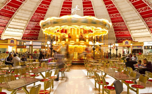 Ein geräumiges Restaurantinnere mit zahlreichen Tischen und Stühlen, das einen Karussell in der Mitte, sitzende und stehende Menschen und eine sichtbare Decke mit Hintergrundbeleuchtung, Beschriftungen und strukturelle Elemente aufweist.
