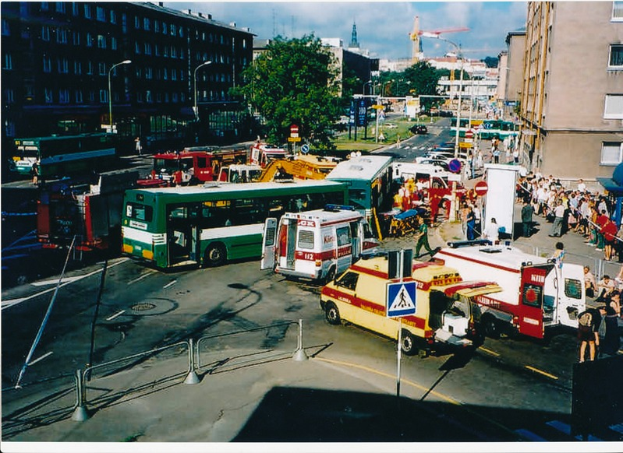 Eine Gruppe von Einsatzfahrzeugen steht auf der Seite einer Straße, umgeben von Menschen, Pfählen, Lampen, Schildern, Bäumen, Gebäuden und Kränen, mit einem bewölkten Himmel im Hintergrund.