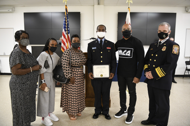 Eine Gruppe maskierter Menschen vor Flaggen, wobei eine Person in der Mitte ein Zertifikat hält, wahrscheinlich bei einer Abschlussfeier.