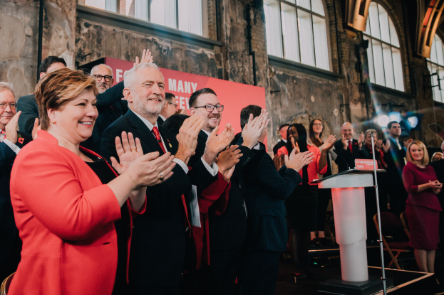 Eine Gruppe von Menschen, wahrscheinlich Liberale, klatscht feierlich vor einem Publikum, mit einem Podium, Mikrofon und Texttafel rechts, Stühlen, einer Fahne, einer Wand, Fenstern und Lichtern im Hintergrund.