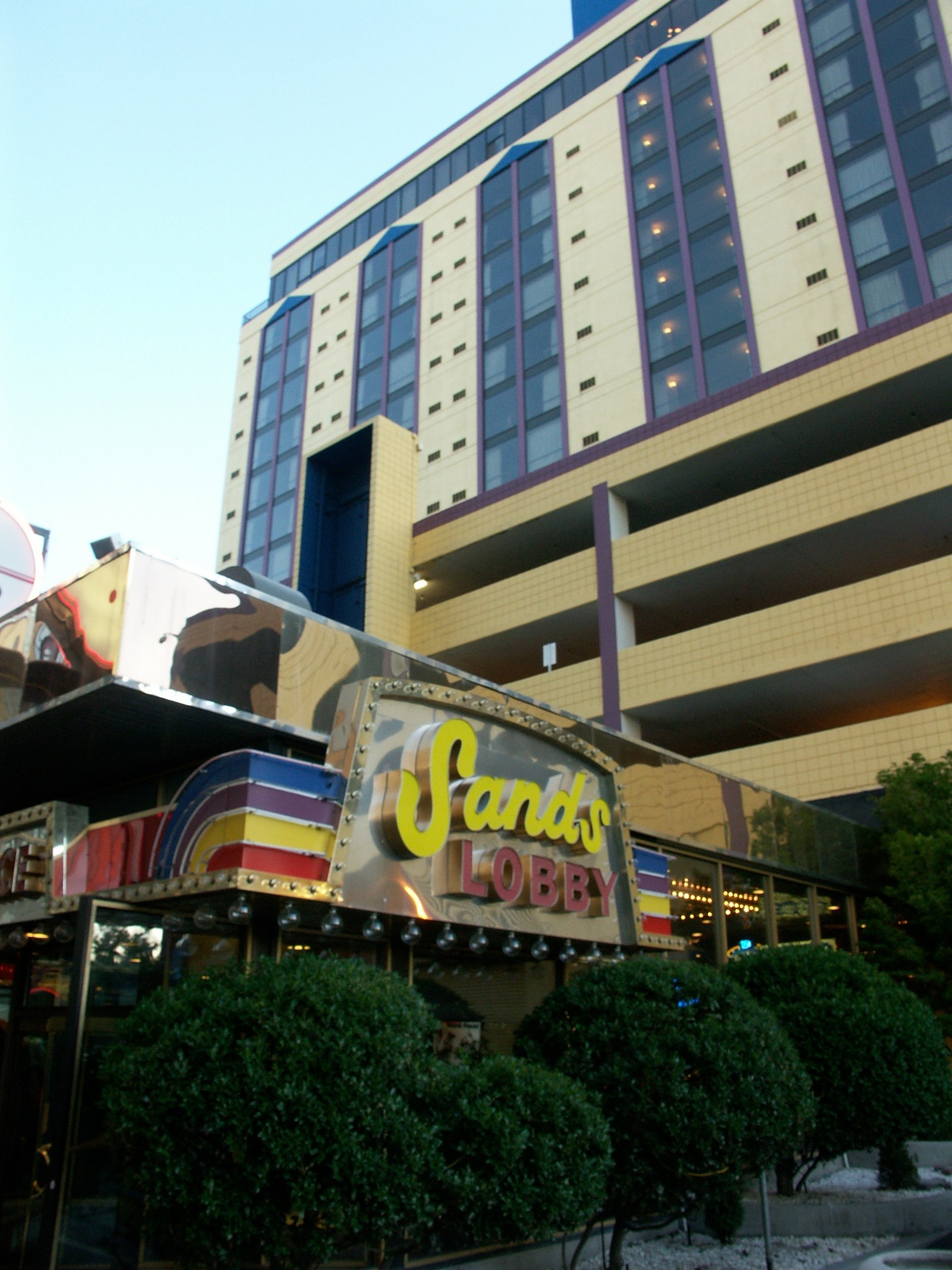 Großes Glasgebäude des Sands Hotel und Casinos in Las Vegas, Nevada, mit einem Namensschild davor, umgeben von Bäumen und Pflanzen unter einem sichtbaren Himmel.