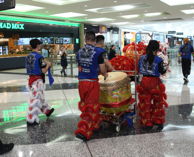 Eine Gruppe von Menschen in roter und blauer Kleidung durchquert einen Flughafen, einige tragen Musikinstrumente und Koffer, mit Geschäften, Namensschildern und Deckenleuchten im Hintergrund, was auf eine chinesische Neujahrsfeier in Singapur hinweist.