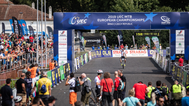 Radfahrer auf einer Straße mit Zuschauern auf beiden Seiten, unter einem Bogen mit der Aufschrift "2019 UEC MTB European Championships", mit Bäumen, Gebäuden und einem klaren blauen Himmel im Hintergrund.