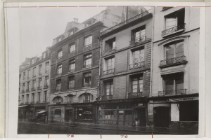 Schwarzes und weißes Foto einer Stadtstraße mit Gebäuden mit Fenstern, Geländern und Namensschildern sowie einer Straße unten.