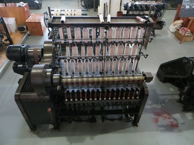 Eine Textilmaschine mit einem großen rechteckigen Rahmen und verschiedenen Knöpfen und Bedienelementen steht in der Mitte eines Raumes, umgeben von anderen Maschinen und Gegenständen, mit einer Tafel mit Text auf der rechten Seite und mehr Maschinen im Hintergrund.