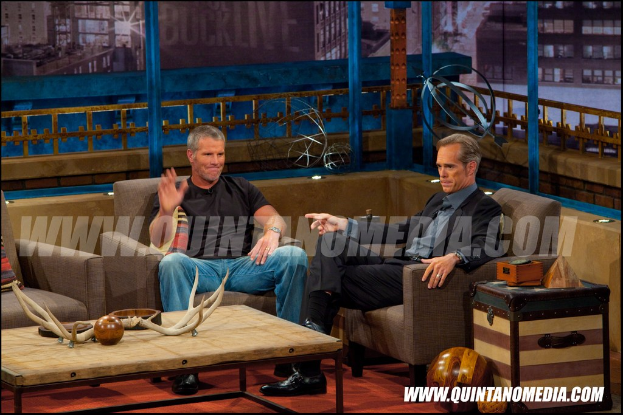 Zwei Personen sitzen auf einem Sofa während eines Medieninterviews, mit einem Tisch und Spielzeugen davor und Wandplakaten dahinter; ein Mann im Blazer hat einen Tisch mit Gegenständen und eine hölzerne Kugel daneben.