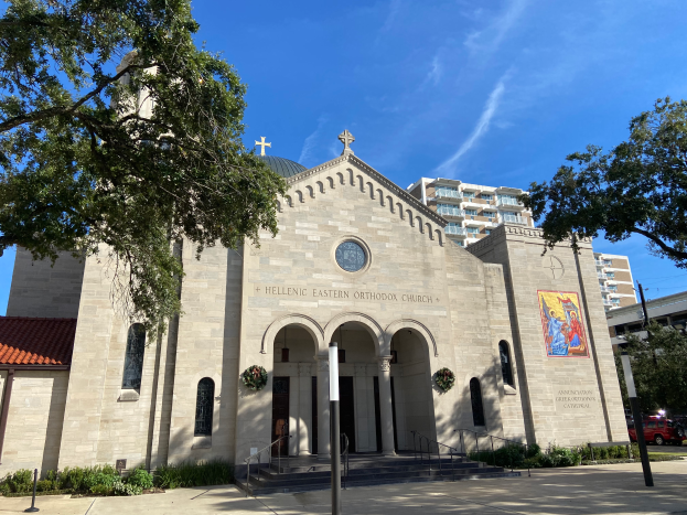 Die griechisch-orthodoxe Kirche in San Antonio, Texas, ist ein großer Bau mit Fenstern, Säulen, Bogengängen und Treppen mit Geländern, umgeben von Pflanzen, Bäumen und Fahrzeugen auf der Straße, unter einem bewölktem Himmel.
