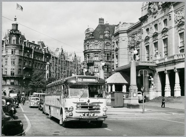Ein Schwarz-Weiß-Foto einer Stadtstraße mit einem Bus, der darauf fährt, umgeben von Gebäuden mit Fenstern, Bäumen, Laternen, einer Fahne mit einem Mast und einigen Menschen.