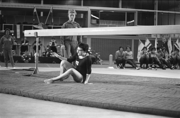 Eine Frau in einem schwarzen und weißen Turnanzug führt eine Übung auf einem Schwebebalken in einer Turnhalle durch, mit Zuschauern und Equipment im Hintergrund.