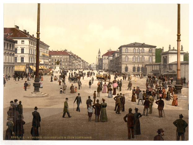 Ein altes Foto einer belebten Münchner Straße in den frühen 1900er Jahren, mit Menschen, die spazieren gehen, einige mit Schirmen, und einem Pferdewagen im Vordergrund, Gebäuden, Bäumen und Pfählen im Hintergrund und Text am unteren Rand des Bildes.