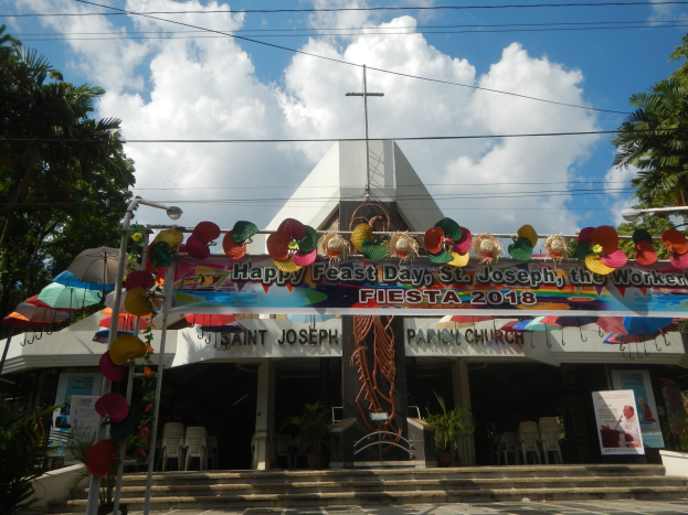 Ein Gebäude mit einer Tafel, die 'Frohes Fest St. Josef der Arbeiter' lautet, davor, mit einer Treppe, die zum Eingang führt, einem dekorativen Bogengiebel, Außenbestuhlung mit Tischen und Stühlen, Topfpflanzen, Bäumen zu beiden Seiten und einem bewölkten Himmel im Hintergrund.