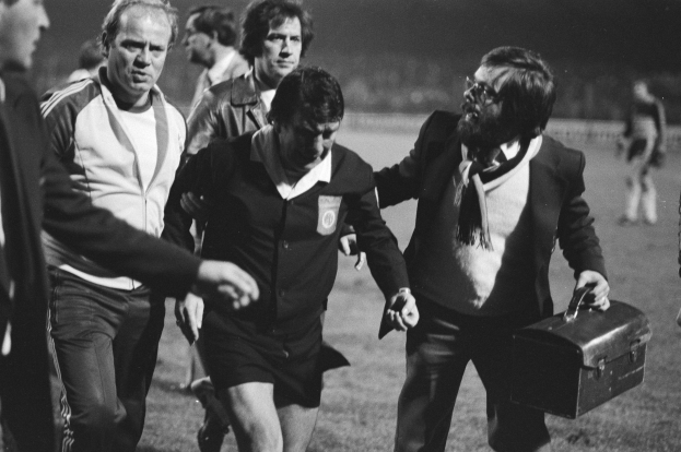 Schwarzes und weißes Bild einer Gruppe von Männern, die auf einem Fußballfeld gehen, einer hält eine Aktentasche und hat einen offensichtlichen Ausdruck von Unbehagen.