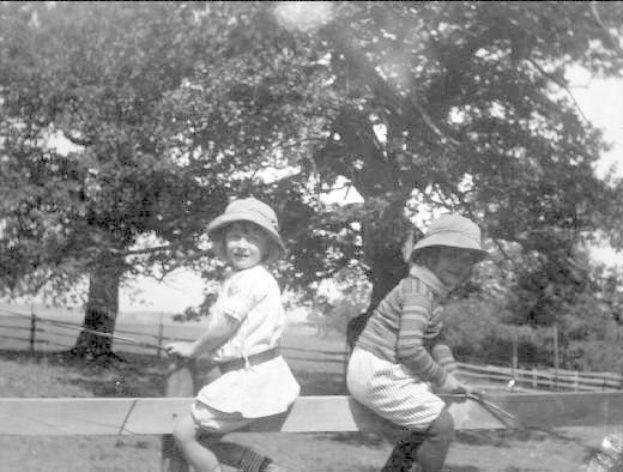 Zwei junge Jungs in Hüten mit Baseballschlägern auf einem grasbewachsenen Park mit Bäumen und einem Zaun im Hintergrund, Schwarz-Weiß-Foto.