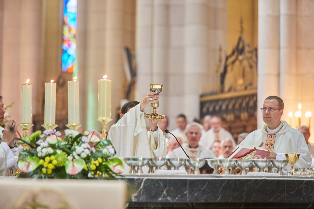 Ein Priester hält einen Kelch vor eine Gemeinde in einer Kirche, mit Gläsern, einem Mikrofon, einem Buch und Blumen im Vordergrund und Kerzen, Säulen und Buntglasfenstern im Hintergrund.