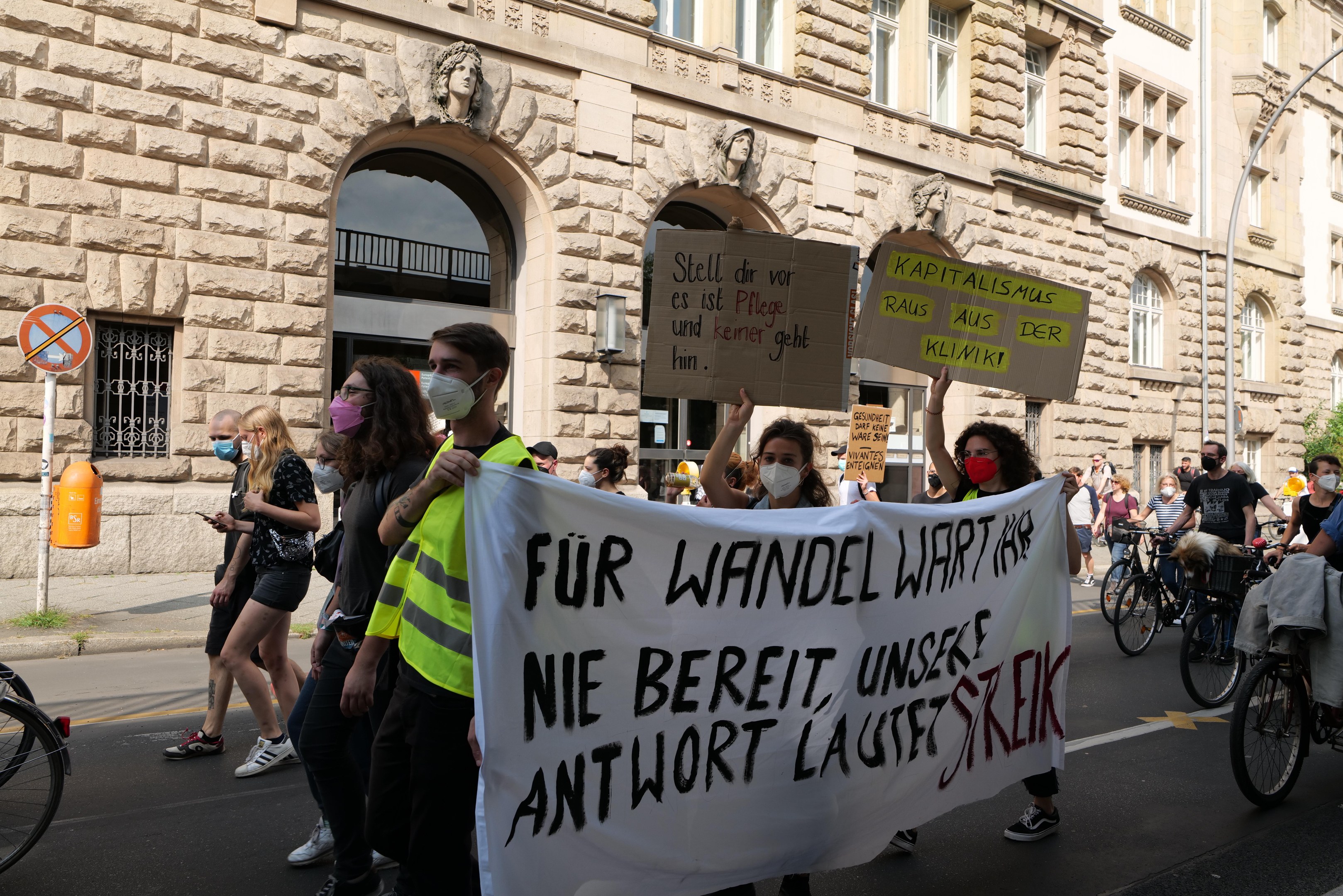 Eine Gruppe von Menschen marschiert durch eine Straße, einige halten Protestschilder und andere fahren Fahrräder, mit einem Gebäude mit Fenstern, Bögen, Säulen und Skulpturen im Hintergrund.