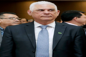 Ein Mann in einem marineblauen Anzug, weißem Hemd und gemustertem Schlips steht vor einer Gruppe von Menschen und hat einen ernsten Gesichtsausdruck und ordentlich frisiertes Haar.