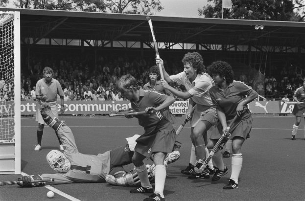 Eine Gruppe von Frauen, die Feldhockey spielen, in Schwarz-Weiß, mit Helmen und Hockeystöcken, mit einem Torpfosten links, Zuschauern auf den Rängen, Bannern, einer Hütte, Bäumen und einer Flagge im Hintergrund.