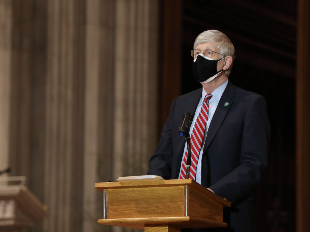 Ein Mann im Anzug und Krawatte trägt eine Gesichtsmaske und steht an einem Podium mit einem Mikrofon und einem Buch, während er eine Rede hält. Im Hintergrund sind Säulen und eine Wand zu sehen, sowie ein weiteres Podium links von ihm.