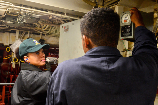 Zwei Personen, ein Mann mit Mütze und eine Frau mit Kopfhörern, bedienen Maschinen in einem Raum voller Geräte und Kabel.
